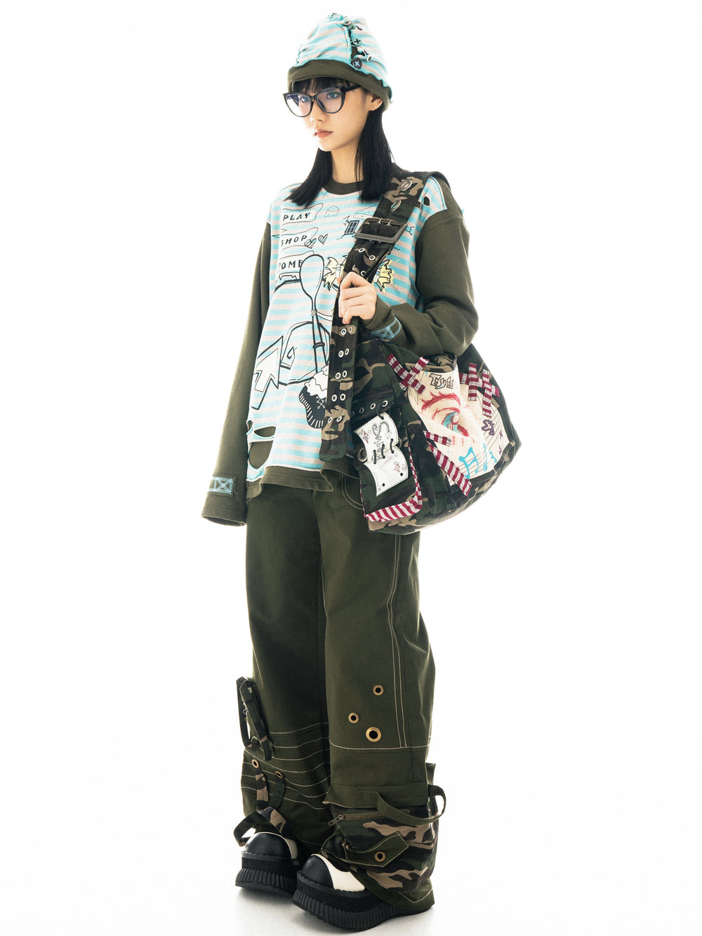 full shot of a female model showcasing a stylish outfit. she's wearing a light blue longsleeved shirt with graphic print, olive green cargo pants with unique detailing, camouflage print boots, and a matching camouflage and patterned bag.  she also sports a light blue beanie and glasses. the background is plain white. the overall style is a blend of streetwear, harajuku, and possibly some japanese fashion influences.