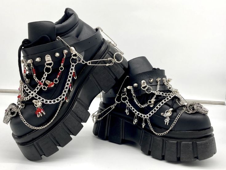 two black platform boots with goth and punk aesthetics are displayed. the boots are adorned with silver chains, skulls, spikes, and safety pins. they have thick soles and a layered design, giving them a distinct and edgy look.