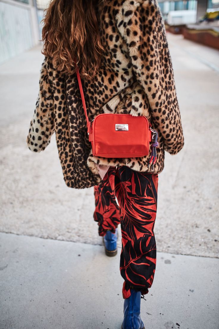 a woman walks down a city street, her back to the camera. she is wearing a leopard print coat, red tropical print pants, and blue doc martens. she carries a red crossbody bag. the image is taken from a slightly low angle, giving a good view of her outfit and accessories. the overall style is trendy and chic.