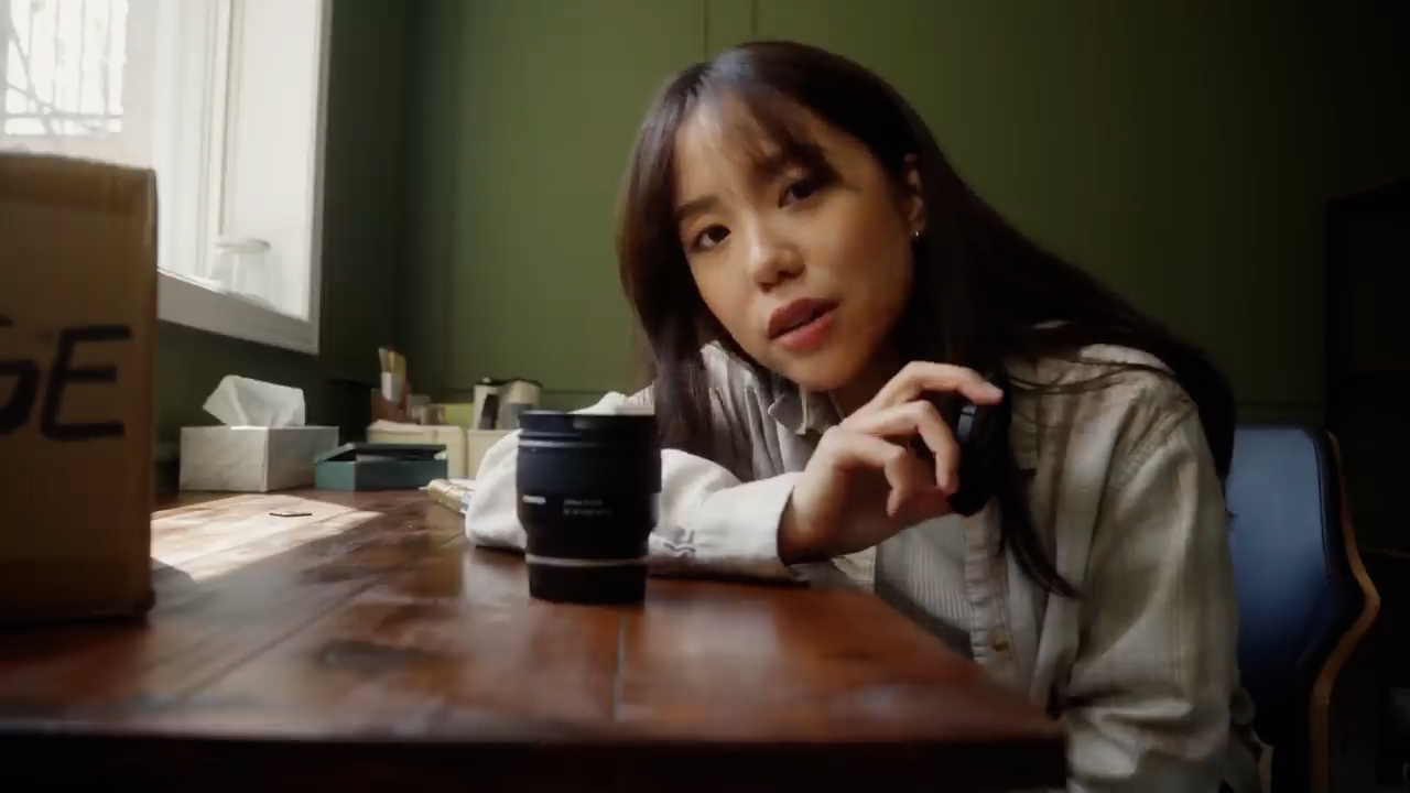 a young woman sits at a wooden table in a naturally lit room. she's holding a camera lens and there's a camera lens box on the table. she seems to be talking to the camera, possibly reviewing the lens. the overall atmosphere is warm and inviting.