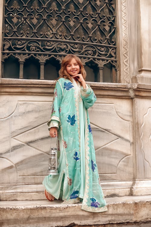 a young girl, possibly of middle eastern descent, stands elegantly in front of an old stone building. she wears a long, flowing teal dress with a floral pattern and ornate detailing.  the dress has a traditional style, possibly a kaftan or abaya. she holds an antiquelooking lantern in one hand. the building's architecture is grand and features an intricately designed iron window grate. the overall aesthetic is stylish, elegant, and evokes a sense of cultural heritage and travel. the girl's pose and smile project confidence and charm. the scene is welllit by natural daylight.