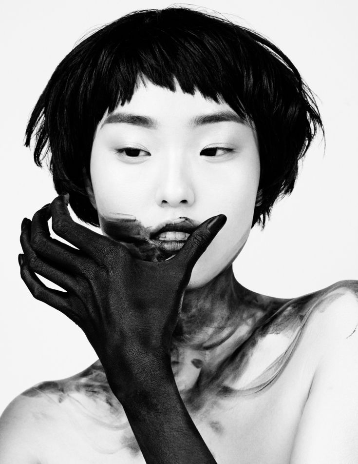 a black and white portrait of a woman with black paint covering her face and hands, with a mysterious and expressive look. the woman's short dark hair and the paint create a dramatic and artistic composition. the photo has a closeup perspective highlighting the woman's features, suggesting a sense of intimacy and vulnerability.  the lighting is soft and even, highlighting the textures of the paint and the skin.  the image is likely part of a fashion or art project, showcasing the woman's beauty and captivating gaze.