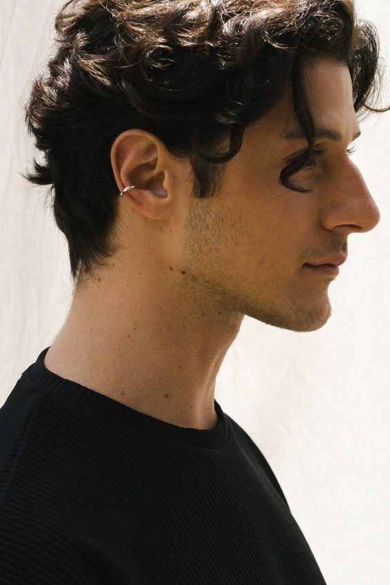 a profile shot of a lightskinned man with curly dark hair. he is wearing a black shirt and has an ear cuff on his left ear. the background is a light offwhite color.