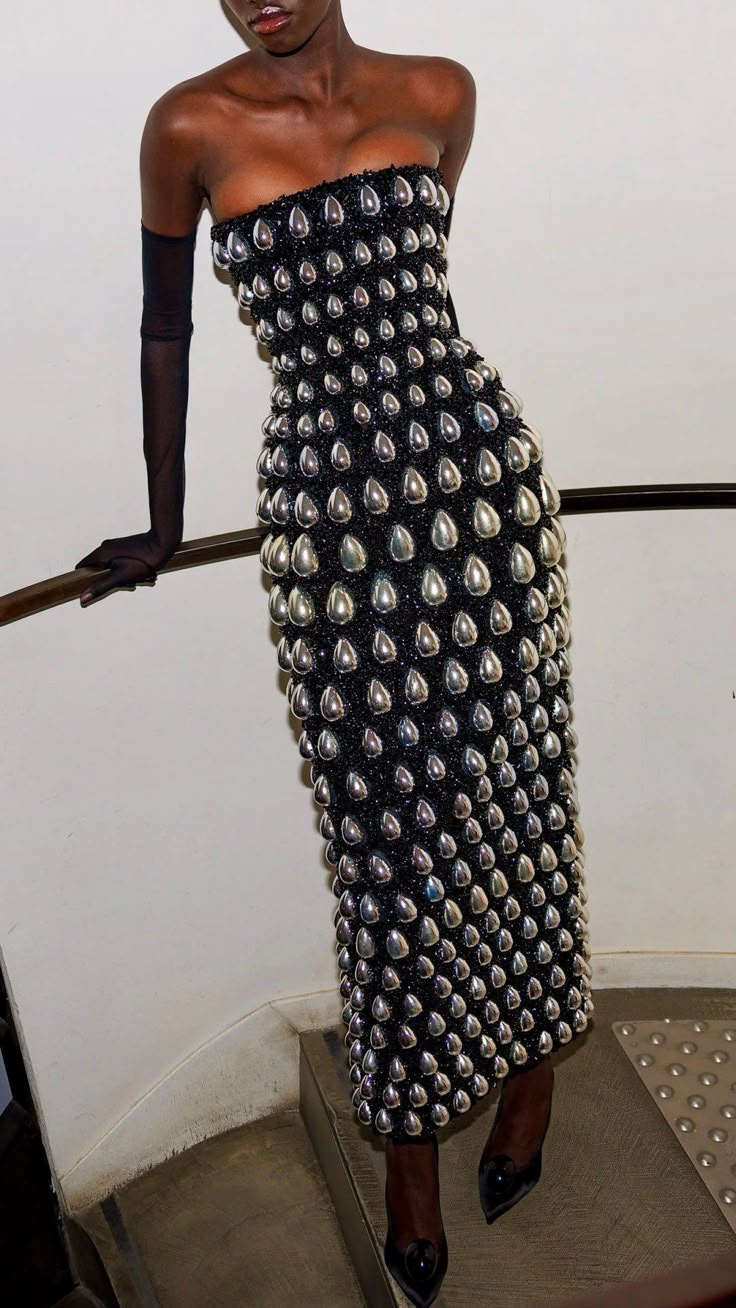 a black woman stands on a set of stairs, leaning against the railing. she is wearing a formfitting, strapless black dress embellished with silver, teardropshaped embellishments. she has on sheer, black opera gloves. she is wearing black high heels. the background consists of white walls and part of the metal stairs.