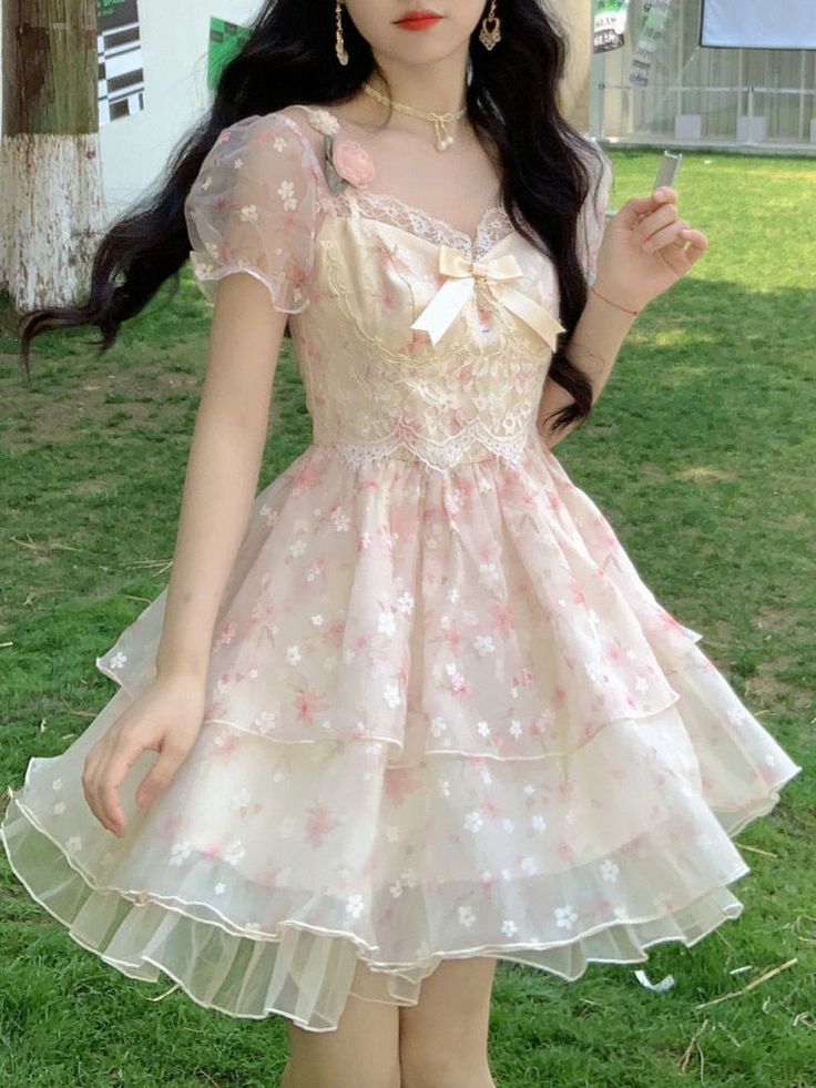a young woman poses for the camera while wearing a flowing, pastel pink dress with a floral pattern. she stands on a grassy area, and the background suggests an outdoor park or public space. the lighting is bright and natural. her dress features puffed sleeves, a sweetheart neckline, a layered skirt, and is decorated with lace detailing and a bow.