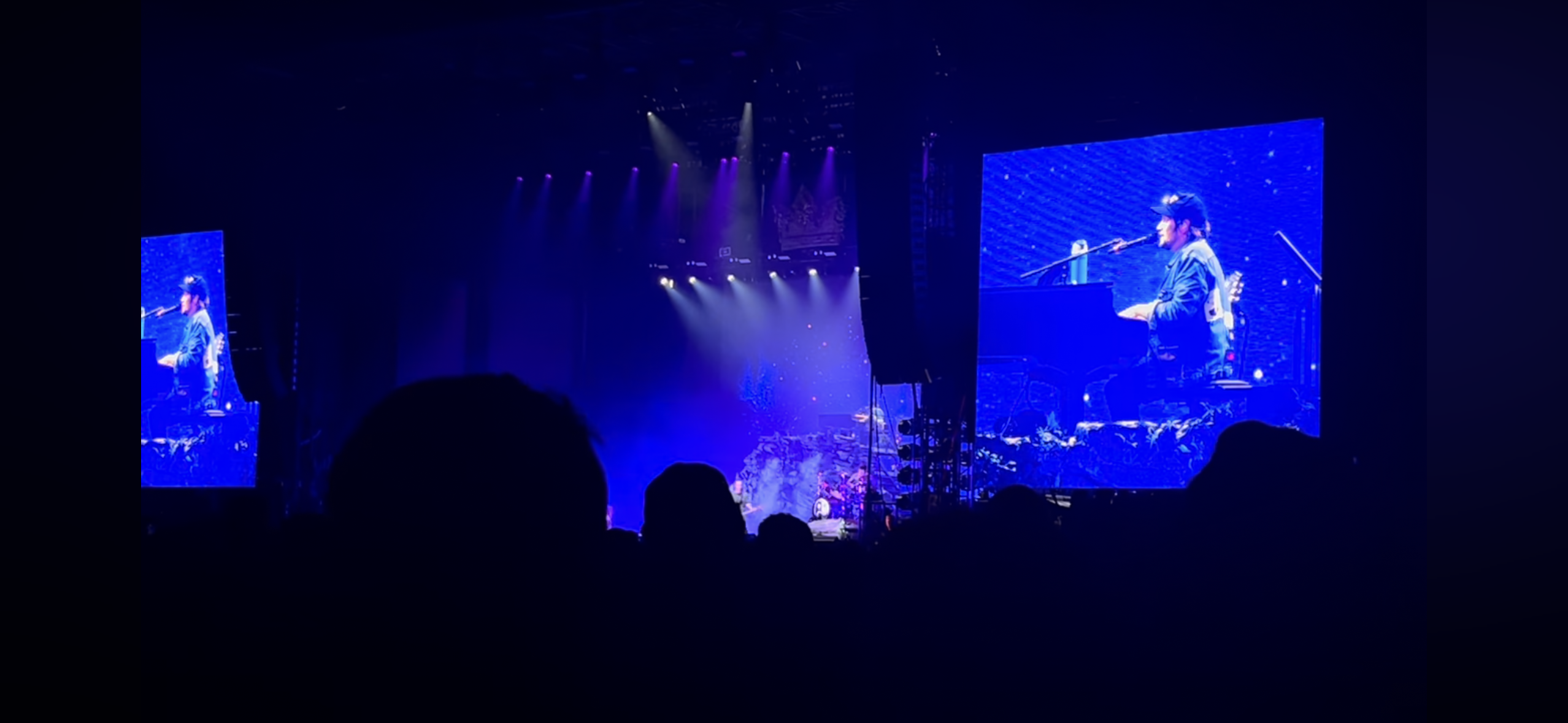 a concert scene with a dark blue ambiance, featuring a performer playing the piano on a large screen. the audience is mostly silhouetted in the foreground, and there are bright blue lights illuminating the stage.