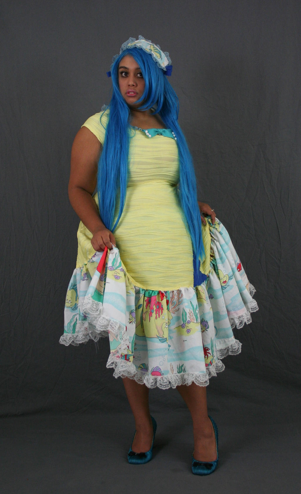 a fullbody shot of a woman with bright blue hair, wearing a pastel yellow dress with a white lace trim and a colorful underlayer. the dress is kneelength. the woman is wearing teal heels. she is standing on a gray background. the woman is looking directly at the camera. she is wearing a small, lightcolored headpiece.