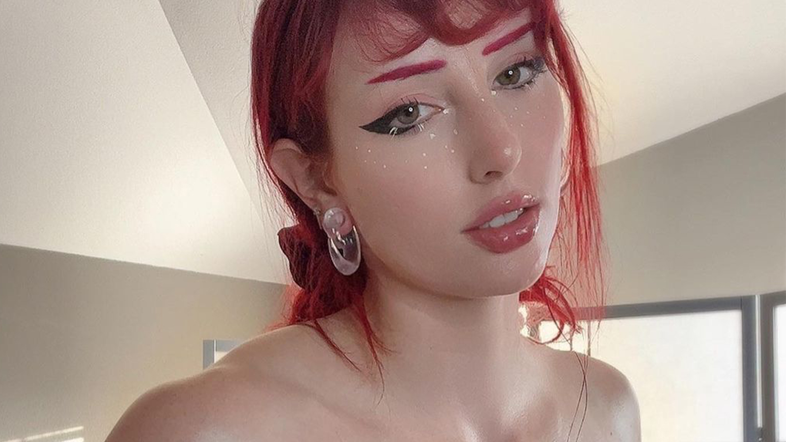 a closeup selfie of a young woman with vibrant red hair. she's wearing striking makeup featuring winged eyeliner, bold red eyebrows, and small, strategically placed freckles.  her earrings are large, clear hoops.  her expression is soft and she's looking slightly offcamera. the background is blurred but shows a portion of a room with beige walls. the overall aesthetic is alternative and stylish.