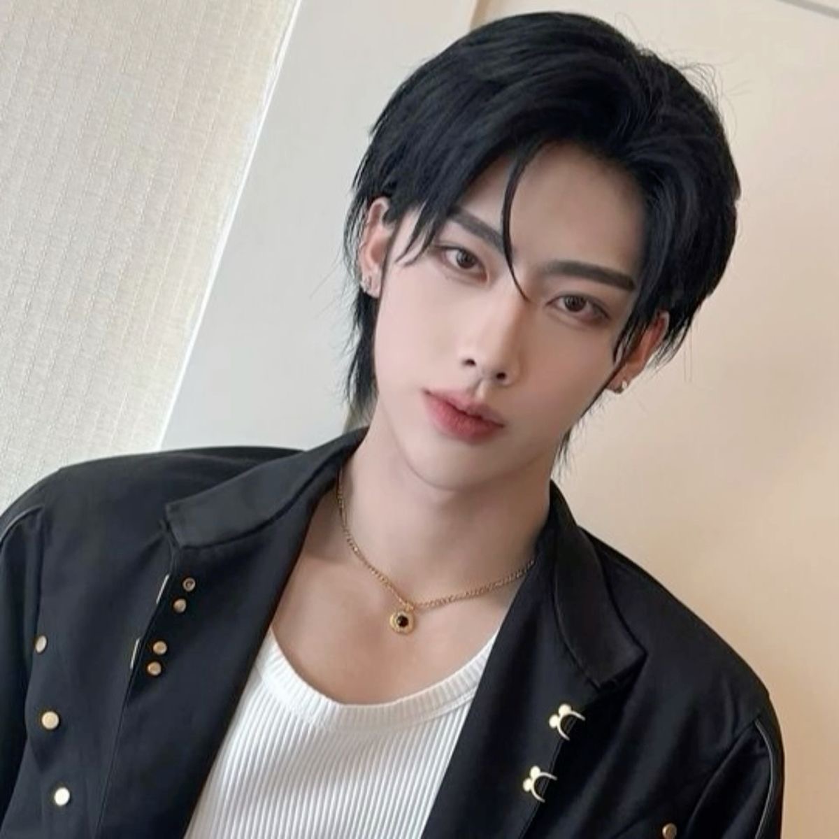 a head and shoulders portrait of a young, androgynous asian man. he has black hair styled with a middle part and long bangs framing his face. he is wearing a black collared jacket with silver stud details and a plain white shirt underneath. he is also wearing a thin gold chain necklace with a round pendant. the background is blurred, but appears to be an indoor space with light walls.  the lighting is artificial, and the image appears to be a selfie.