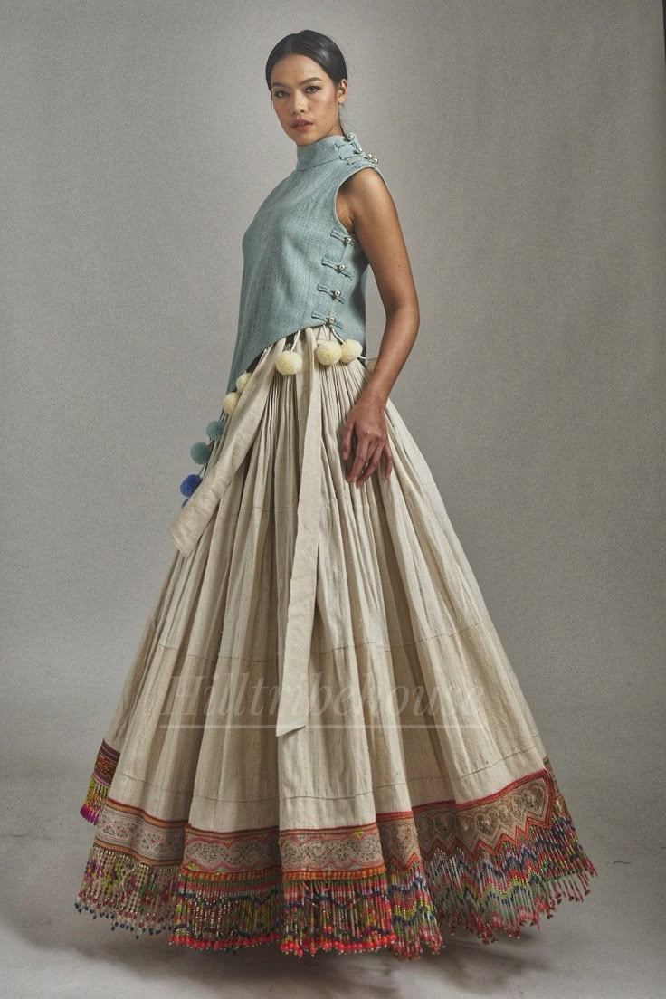 a woman is standing in a studio setting, wearing a unique and ornate ethnicinspired outfit. she has a pale blue, sleeveless, highnecked top with small, buttonlike closures down one side. the skirt is a full, flowing design in an offwhite color, cinched at the waist with a tie, and decorated with large, fluffy pompoms around the waistband. the hem of the skirt features intricate embroidery with zigzag patterns and colorful beaded trim. the overall look is a blend of traditional elements with a modern fashion sensibility.