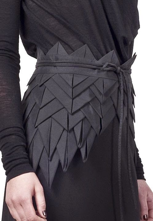 a closeup shot of a woman wearing a black dress with a unique origamiinspired textured belt. the dress has long sleeves and a slightly draped top. the belt is the focal point, featuring a series of folded, geometric shapes reminiscent of origami. it's tied at the side with a thin cord, adding to the artistic and unconventional style.