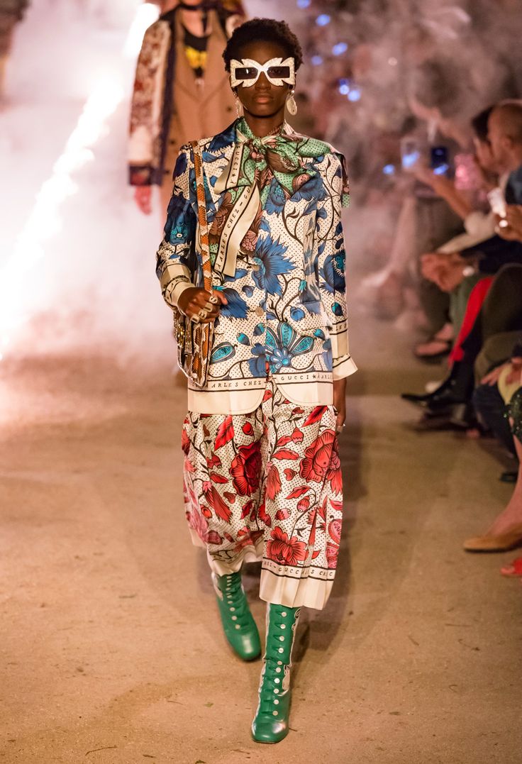 a model walks the runway at a gucci fashion show. she is wearing a striking ensemble featuring a floralprinted silk jacket and pants, paired with green laceup boots and white sunglasses. the background is blurred, showing other attendees and a smoky atmosphere. the overall scene is one of high fashion and luxury.