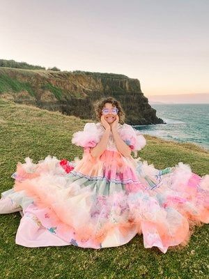 a smiling woman with curly hair and glasses is sitting on a grassy area near a cliff overlooking the ocean. she is wearing a colorful, elaborate gown. her hands are gently placed on her face, as if in awe or delight. the background shows the coastline with a cliff and the ocean.