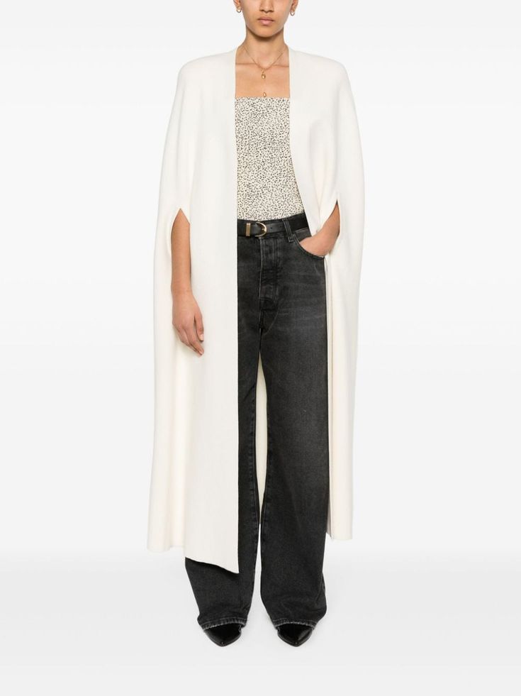 a woman is standing in a white cape, black jeans, black heels, a floral print top, and a gold necklace. she is looking straight ahead.