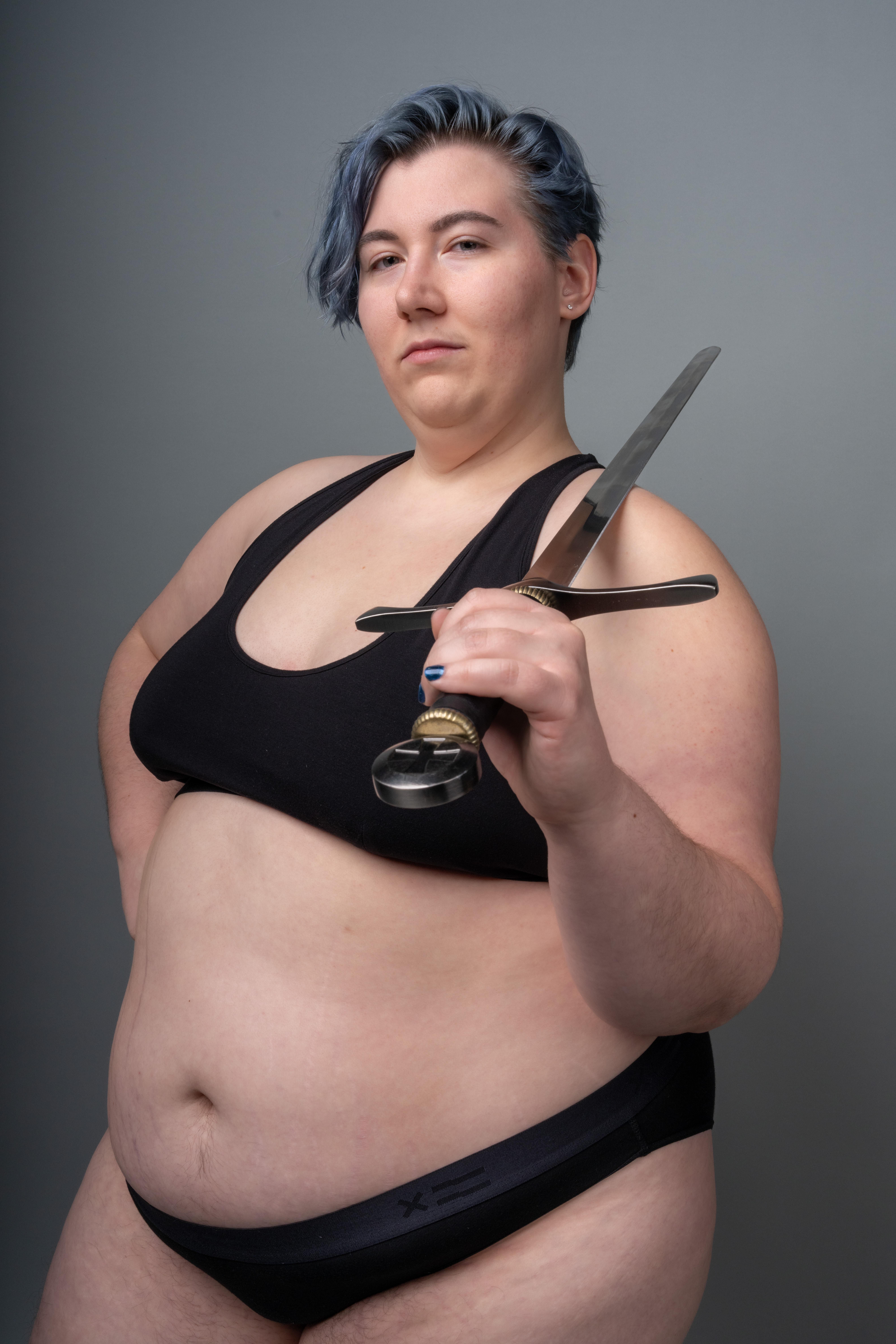 a confidentlooking plussize woman with short blue hair stands against a grey background, holding a sword. she is wearing a black sports bra and black underwear. she looks directly at the camera with a determined expression. the image promotes body positivity and portrays the woman as a strong and selfassured female figure.