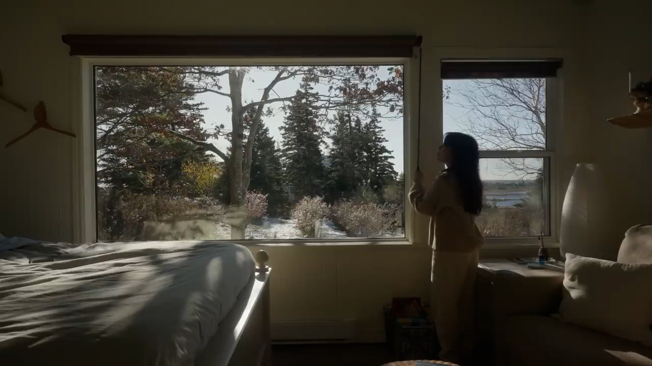 a woman stands by a large window, her back to the camera, gazing out at a sunlit autumnal landscape. the room is a bedroom, with a bed visible on the left side of the image. the sunlight streams through the window, casting warm light on the woman and the room. the overall atmosphere is one of peace and tranquility.
