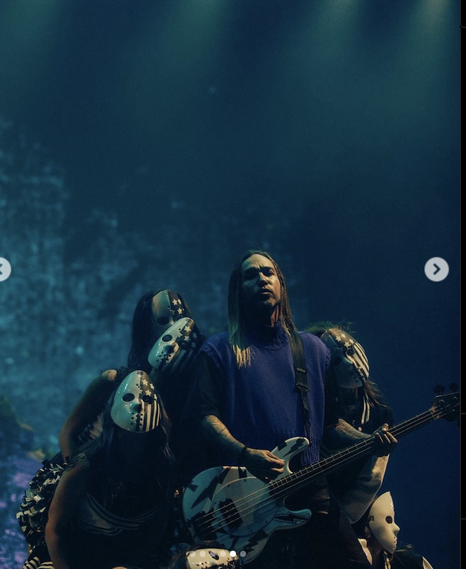 a musician wearing a blue sweater and playing a bass guitar in front of a crowd of people. the musician is looking up at the crowd and smiling. the crowd is cheering and singing along to the music. the atmosphere is electric and exciting. the stage is lit up with bright lights. the musician is having a great time performing. the music is loud and the energy is high.