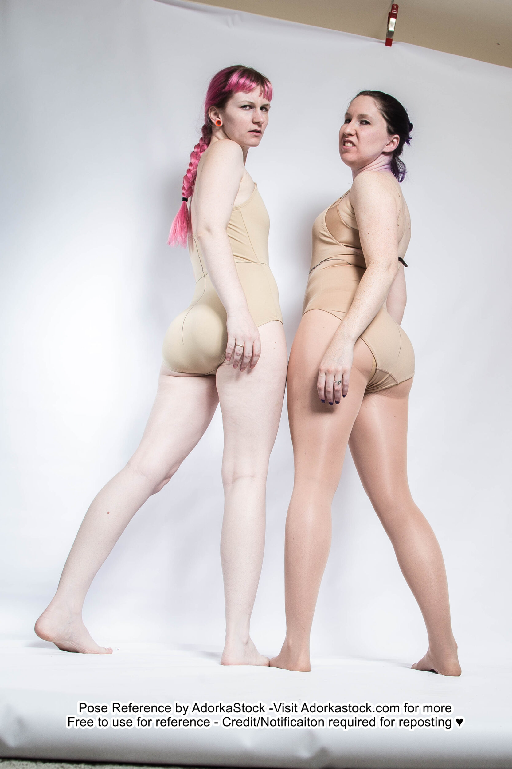 two young caucasian women in beige onepiece swimsuits are posing against a white backdrop. they are facing the camera and have a serious expression on their faces. the woman on the left has pink hair, while the woman on the right has dark hair. the image is a good reference for artists and photographers who are looking to create their own artwork.
