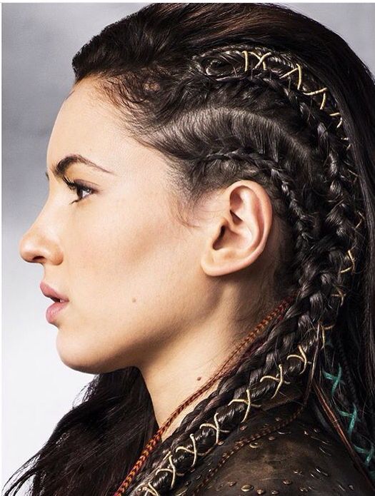 a profile shot of a woman with dark hair styled in multiple braids adorned with goldcolored crisscross wire accents. the braids are arranged along the side of her head, near her ear. she has fair skin, and her facial features such as her nose, lips, and eyebrows are clearly visible. she is wearing a dark garment with studded detailing around the neckline.