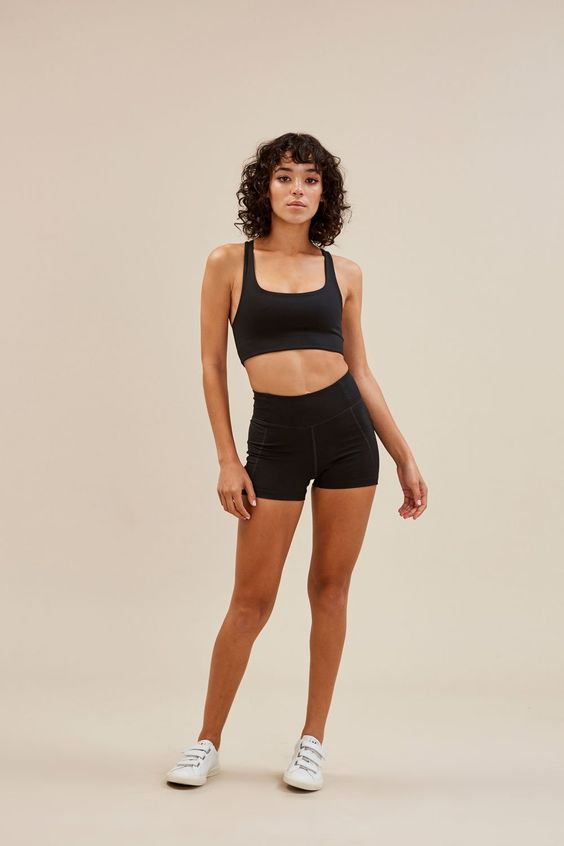 a woman with curly hair is standing against a beige background. she is wearing a black sports bra and black athletic shorts. she is also wearing white sneakers. she is posing with her hands at her sides and her feet slightly apart. she has a neutral expression on her face.