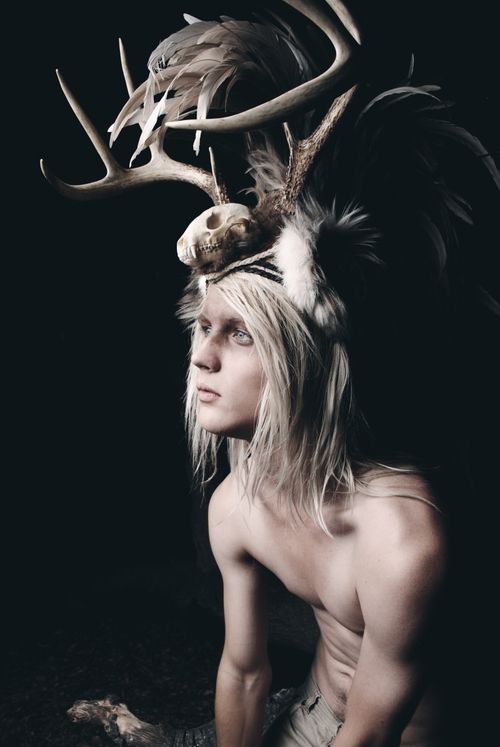 a young man with long white blonde hair is wearing a dramatic headpiece made of antlers, feathers, and a skull. he is barechested and is looking away from the camera. the background is black and the lighting is dramatic.