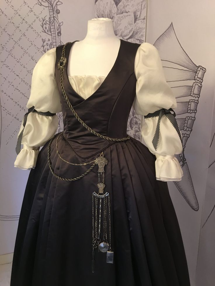 a dress on a mannequin is shown in the picture. the dress has a dark brown bodice with a vneck over a white shirt. the sleeves of the shirt are large, puffy, and white, with gray ribbons at the cuffs. a long, dark skirt descends from the waist. an intricate gold chain runs across the dress, adorned with pendants and trinkets, adding detail to the garment.