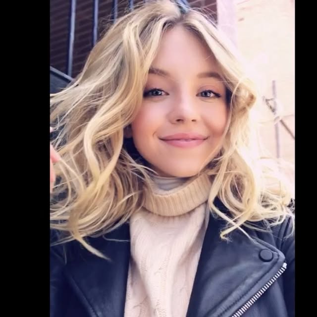 the image is a closeup, slightly lowangle outdoor shot of a young, attractive blonde woman with wavy shoulderlength hair.  she is smiling softly and appears happy. she's wearing a beige turtleneck sweater under a darkcolored leather jacket. the background is slightly blurred but shows a city setting with what looks like a brick building and a metal fence. the overall mood is casual and natural. the lighting appears to be natural daylight.