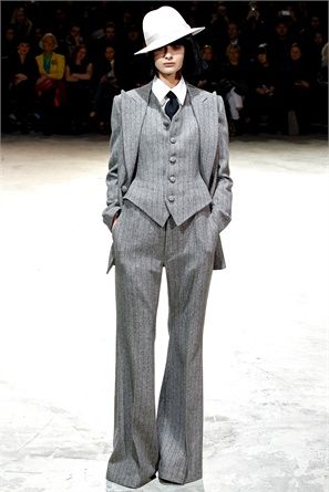 a woman in a grey suit and white hat poses for the camera on a runway. she is wearing a grey vest, white shirt, and black tie. her pants are widelegged and have a pinstripe pattern. her hat is tilted at a jaunty angle, and she is looking straight ahead. the background is blurred, and the floor is white.