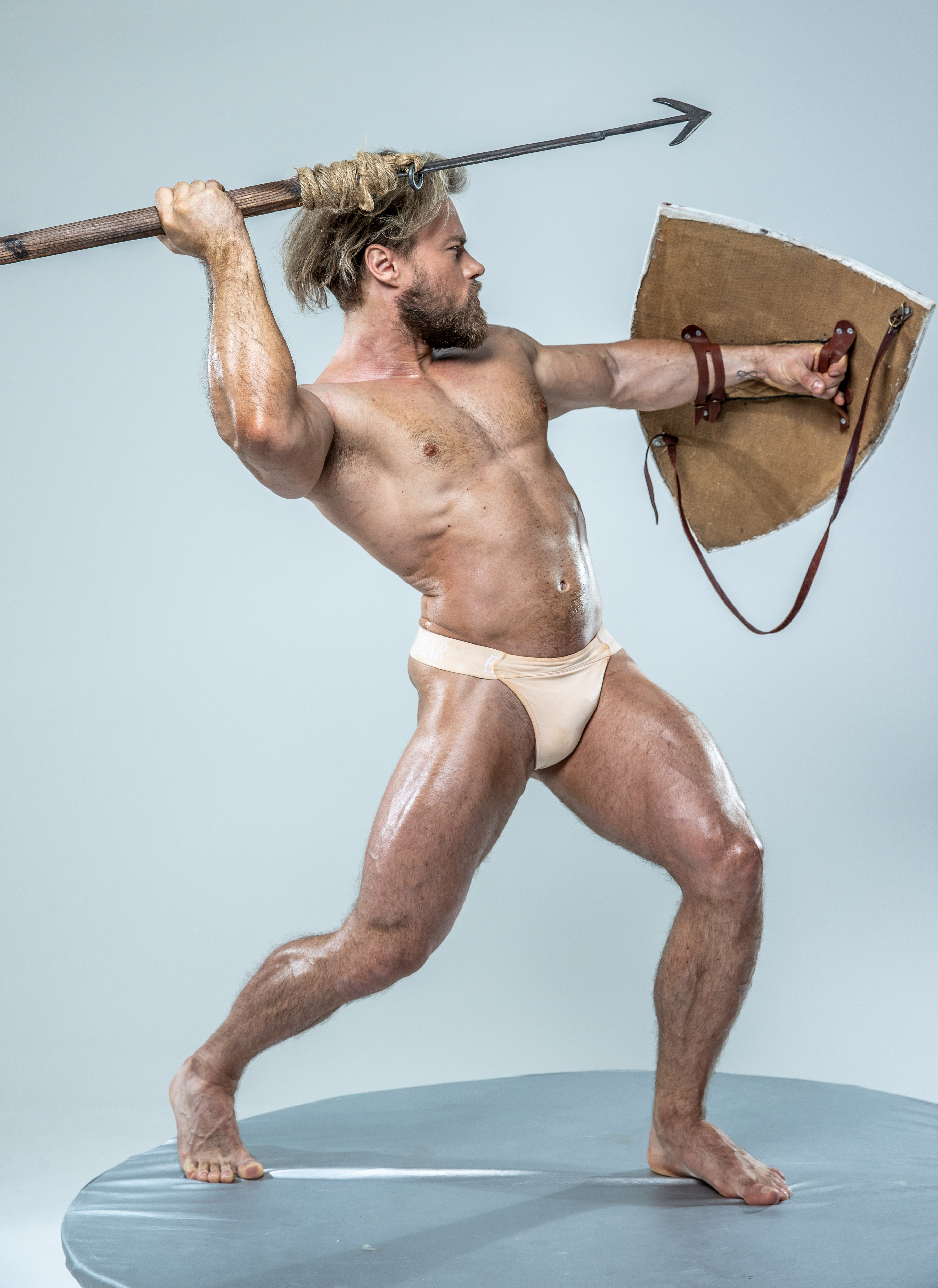 a muscular man with long blond hair, styled in a warriorlike appearance, poses dynamically against a light gray background. he wields a spear with his right hand, poised for an attack, while his left hand holds a round shield, suggesting defense. his body is glistening, implying movement and exertion. the image evokes a sense of power, strength, and the spirit of a warrior. the neutral background and soft lighting emphasize the man's physique and the intensity of his pose.
