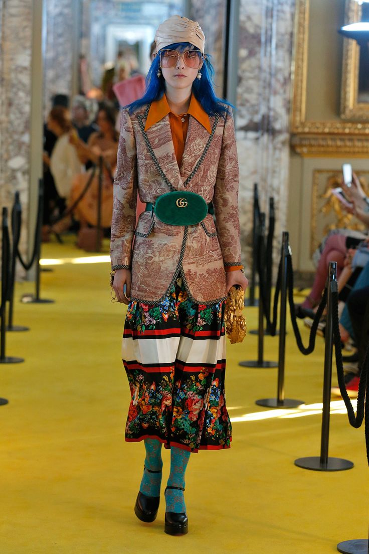a model walks the runway at a gucci fashion show. she is wearing a unique outfit that includes a patterned blazer, a colorful floral skirt, a green gucci belt bag, sunglasses, and a turban. her hair is bright blue. the runway is yellow, and the background features ornate architecture and other people watching the show.
