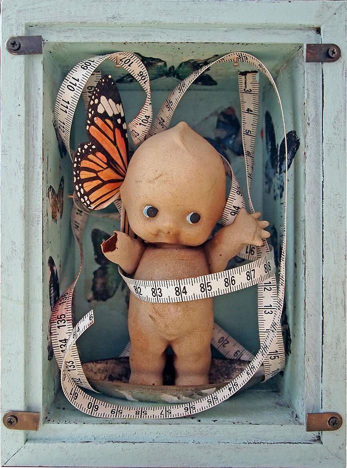 the image shows a mixed media assemblage art piece. a vintage doll stands inside a bluepainted box. the doll has a broken arm and one side adorned with a monarch butterfly wing. a measuring tape is wrapped around the doll and looped within the box. the background of the box is decorated with butterfly imagery. the overall tone is surreal and evocative.