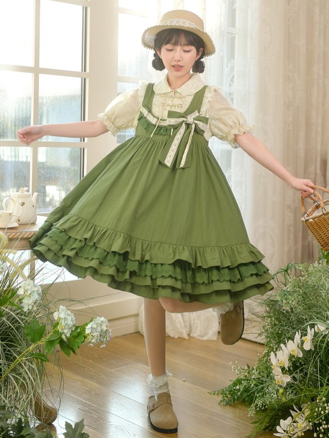 a young woman wearing a green lolita fashion dress poses in a sunlit room. she is holding a straw basket in one hand and her other hand rests on her hip. the dress is a vintage style with lace accents, a layered skirt, and a straw belt. the room is decorated in a cottagecore style with lots of plants and natural light.