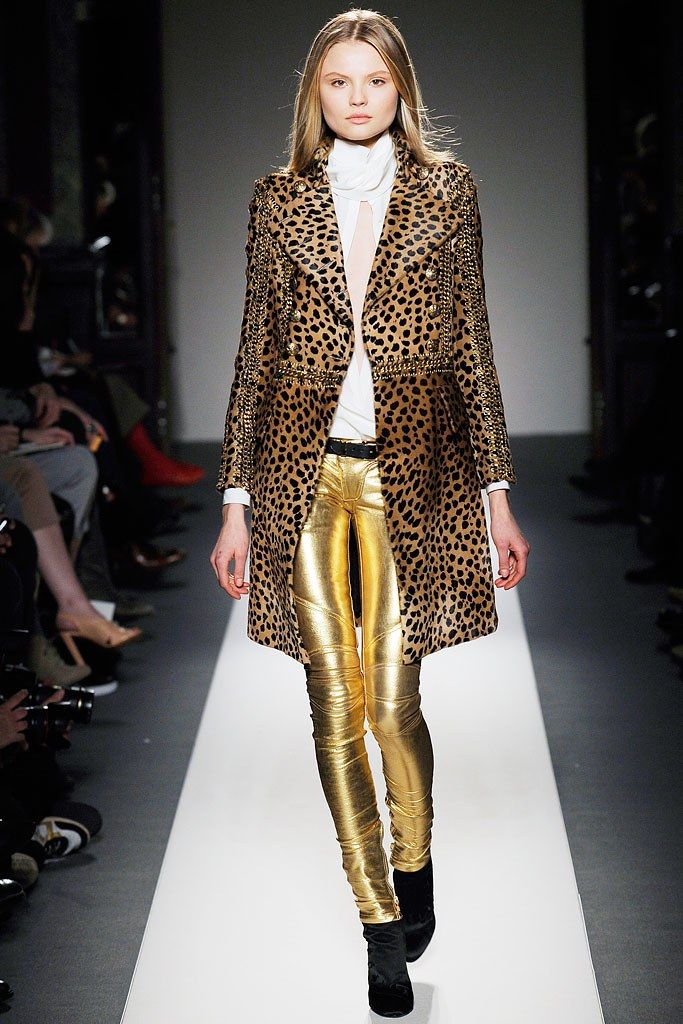 a female model walks the runway wearing a long leopard print coat, gold leggings, and black shoes. she is looking directly at the camera and has a soft smile. the model is wearing jewelry and accessories, including a white shirt underneath her coat. the background is a white runway with a blurred audience.  the model is  wearing a fashionable outfit and is posing confidently. the image is welllit and in focus.
