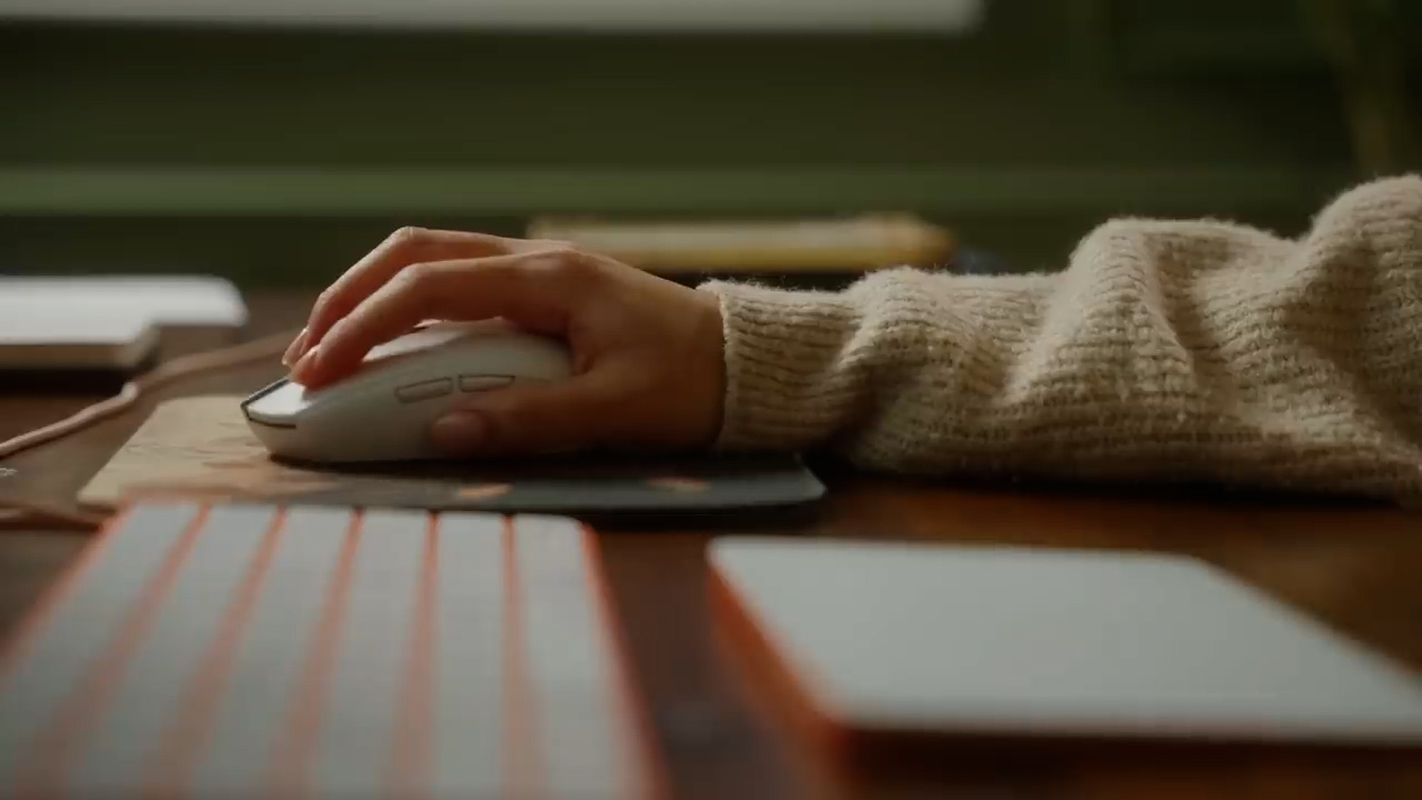 a closeup image of a person's hand using a white computer mouse on a brown desk mat. the image focuses on the hand and mouse, with the rest of the scene, including a keyboard and notebook, slightly blurred. the person is wearing a beige sweater and the lighting suggests a cozy, indoor setting.