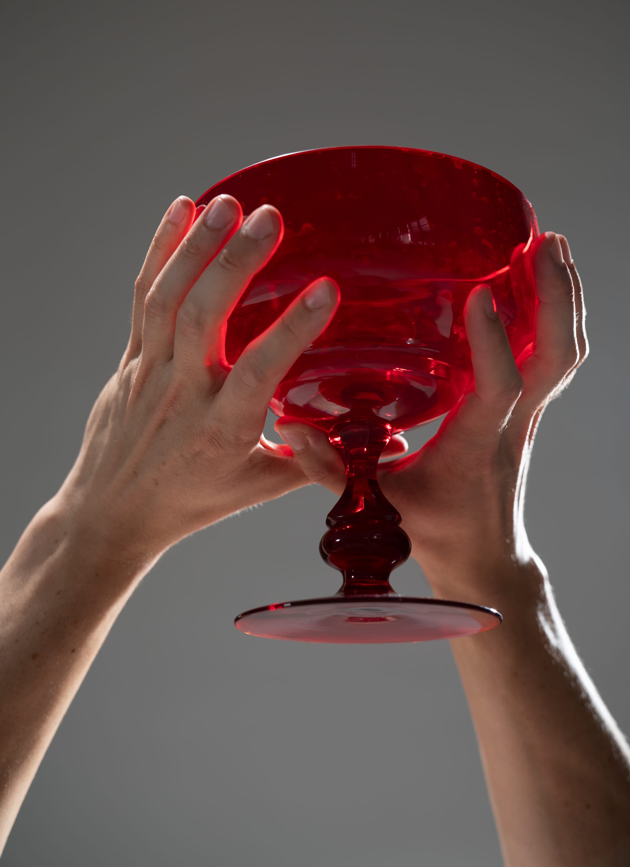 a person holding up a red glass goblet. the goblet is illuminated from below, creating a dramatic effect. the background is a neutral gray.