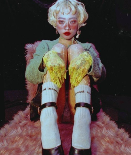 a portrait of a blonde woman sitting on a pink fur chair wearing a green sweater, yellow lace gloves, white socks with black straps, and black mary jane shoes. she is looking directly at the camera with a soft smile and a light makeup. the overall look is very retro and stylish with a soft vintage aesthetic.