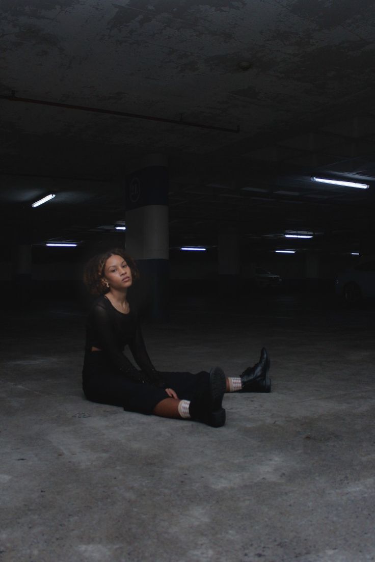 a young woman in all black, sitting on the floor of an underground parking garage with her legs extended forward and her arms resting on her knees. the woman has short, dark hair and a determined expression on her face. she is wearing a black shirt, black pants, and black boots. the floor of the parking garage is grey and concrete.  the image is dark and shadowy, creating a moody and atmospheric feel.  the overall feeling of the image is one of strength and confidence.