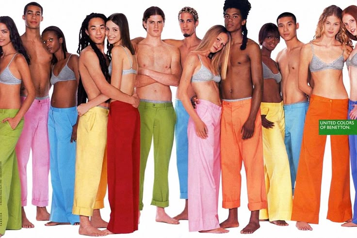a vintage united colors of benetton advertisement featuring a diverse group of young adults. they are standing together, mostly barechested except for bra tops, and wearing brightly colored, widelegged pants in shades of green, pink, blue, yellow, red, and orange. the image aims to convey a message of unity and diversity. the "united colors of benetton" logo is visible in the corner.