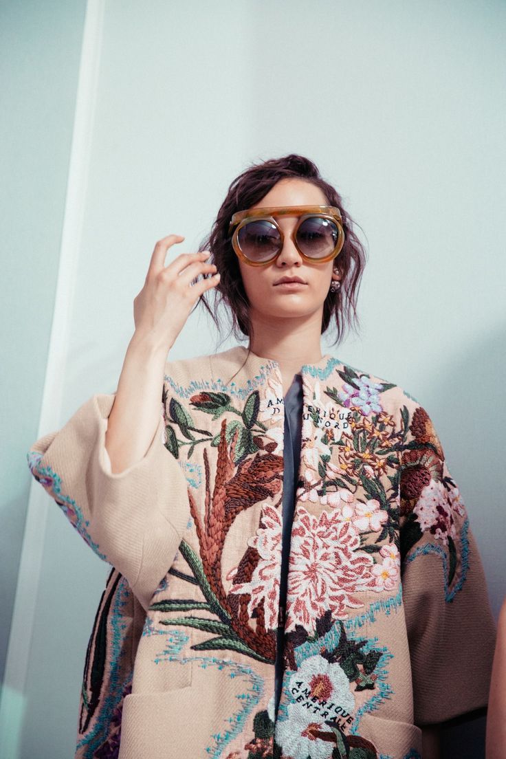 closeup view of a fashion model wearing a beige coat adorned with intricate floral embroidery. the coat features a detailed design with various flowers, leaves, and possibly birds, all embroidered on the fabric.  the model is wearing large, round, retrostyle sunglasses and has dark hair.  the background is a plain, lightcolored wall. the overall style suggests a highfashion, artistic, or possibly vintage aesthetic.
