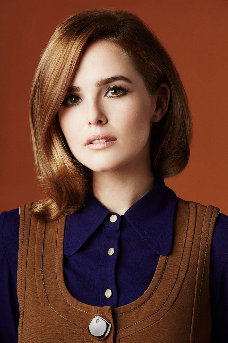 a woman with long brown hair wearing a blue shirt and a brown vest looks directly at the camera. her hair is parted on the side, and she is wearing red lipstick. she has on minimal makeup and is looking directly at the camera.