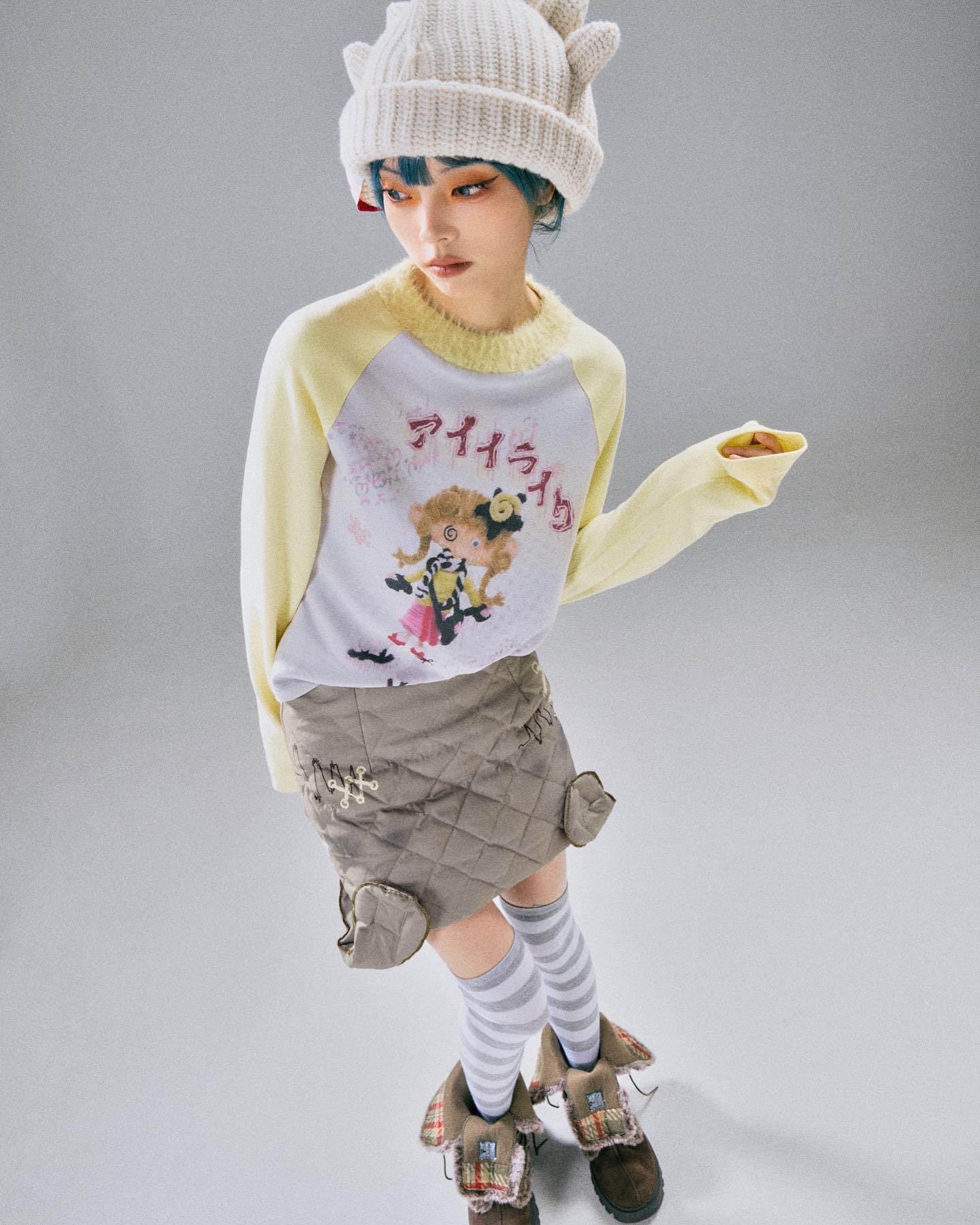 a young woman model is showcasing a stylish outfit. she's wearing a creamcolored knit beanie with small hornlike details on top, a pastel yellow and white raglan longsleeve sweater featuring a cute animestyle graphic print, a quilted beige mini skirt with heartshaped details and subtle embroidery, grey and white striped socks, and chunky brown boots with plaid accents. the overall style is whimsical, blending elements of kawaii japanese fashion with a comfortable, slightly grunge aesthetic. the background is a simple light grey, focusing attention on the outfit and model's pose. the model appears to be looking downwards and slightly to her side.