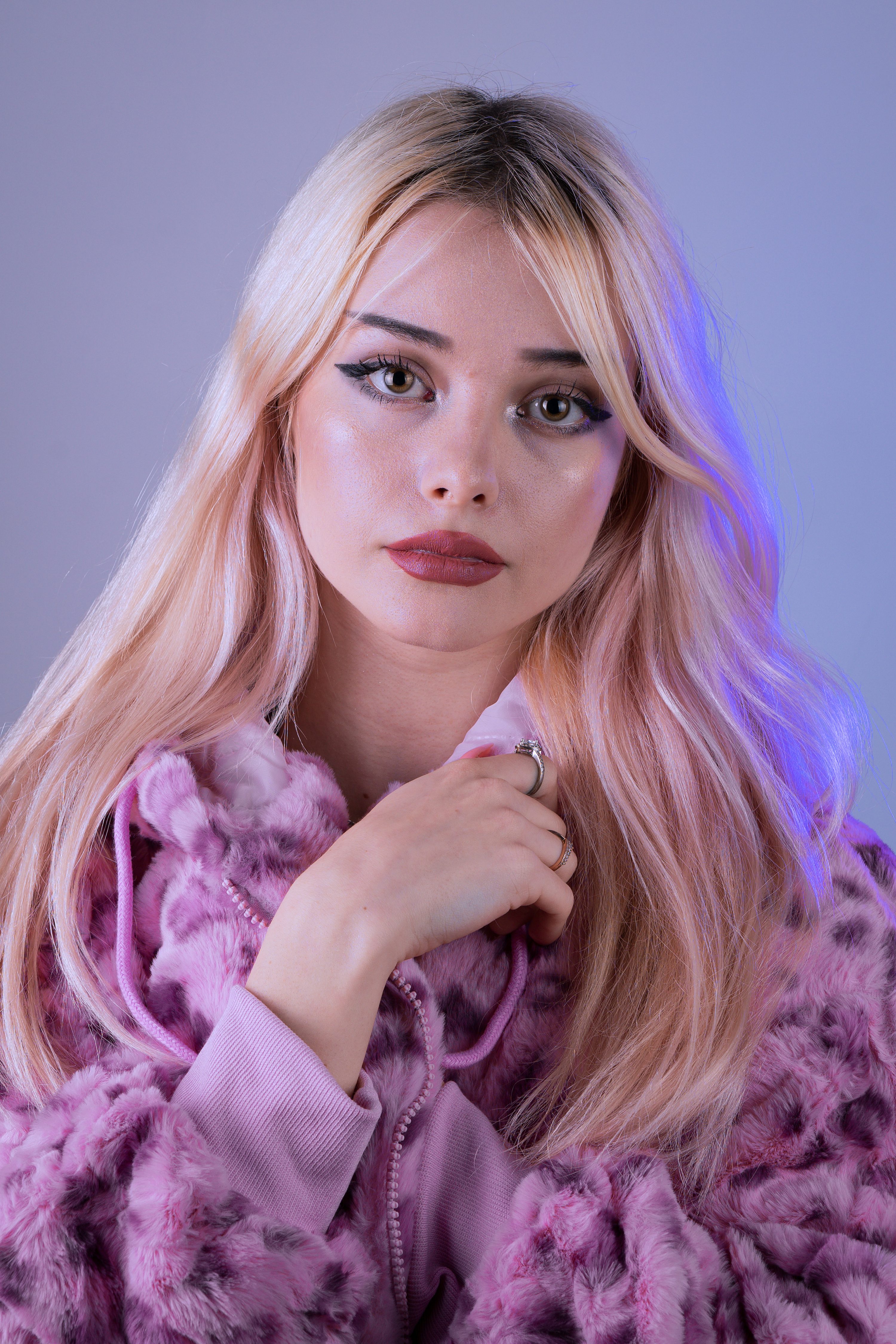 loseup portrait of a young blonde woman wearing a fluffy pink coat with a hoodie. she has long, wavy hair and is wearing subtle makeup. the background is a light purple. the overall mood of the image is soft and gentle.