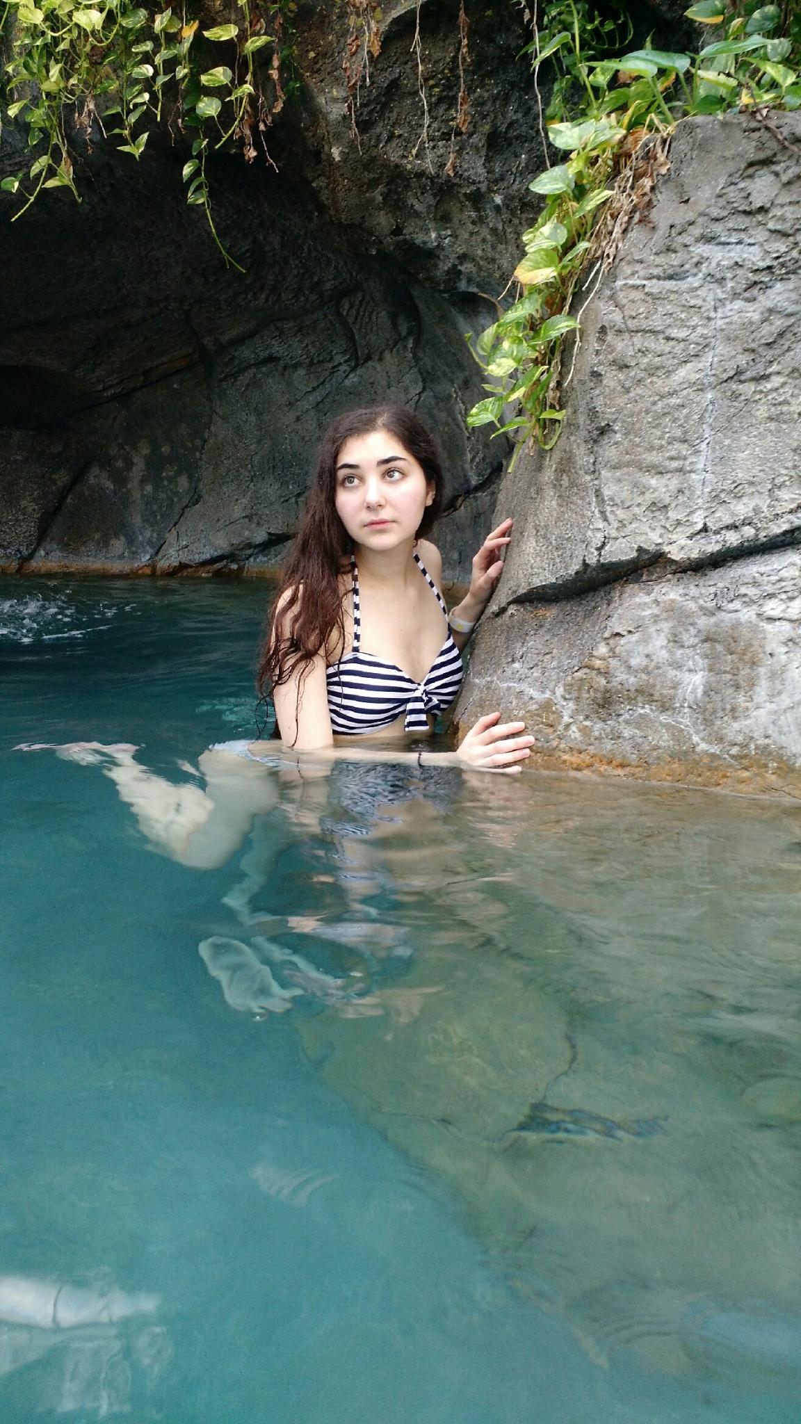 a young woman with long brown hair is partially submerged in a clear turquoise pool of water. she is wearing a black and white striped bikini top. the pool is nestled in a rocky cavelike area, with greenery growing on the rocks. the woman appears relaxed and serene. the overall atmosphere is peaceful and tranquil.
