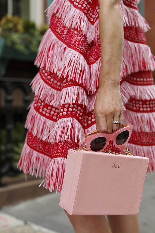 loseup view of a woman wearing a vibrant red and pink fringed dress. the dress is layered with multiple rows of fringe, creating a textured and visually striking effect.  she's carrying a light pink boxshaped handbag with a gold clasp. a pair of pink sunglasses is resting on the top of the handbag.  the background is blurred but shows a city street scene. the overall style is chic and fashionable, suggesting summer style.