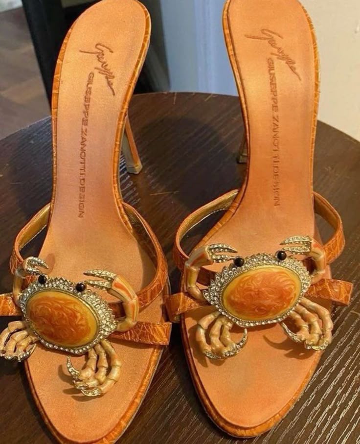 a pair of orange giuseppe zanotti high heels are showcased. the focal point is the crab embellishment on the front strap of each shoe, featuring a jeweled surround and realistic crab details. the shoes have a textured pattern on the upper, likely crocodile or alligator print, and the brand name is visible on the insole. the heels are slender, and the shoes are placed on a dark wooden surface.