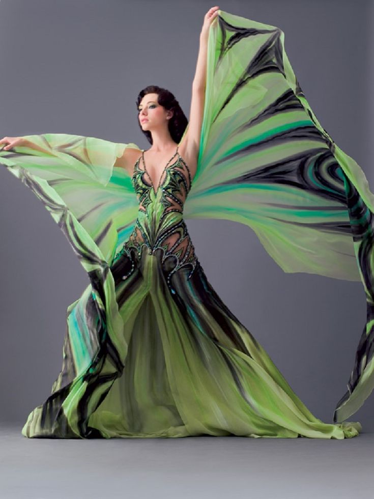 a woman wearing a green dress poses with her arms extended in a dramatic fashion.  the fabric of the dress is flowing behind her, creating a stunning visual effect. she is wearing a beautiful dress with elegant design and a green color that symbolizes elegance and glamour. her pose is graceful and sophisticated. the backdrop is a neutral grey, which allows the dress and the model to be the focal point of the image. the picture looks like an art photo shoot.