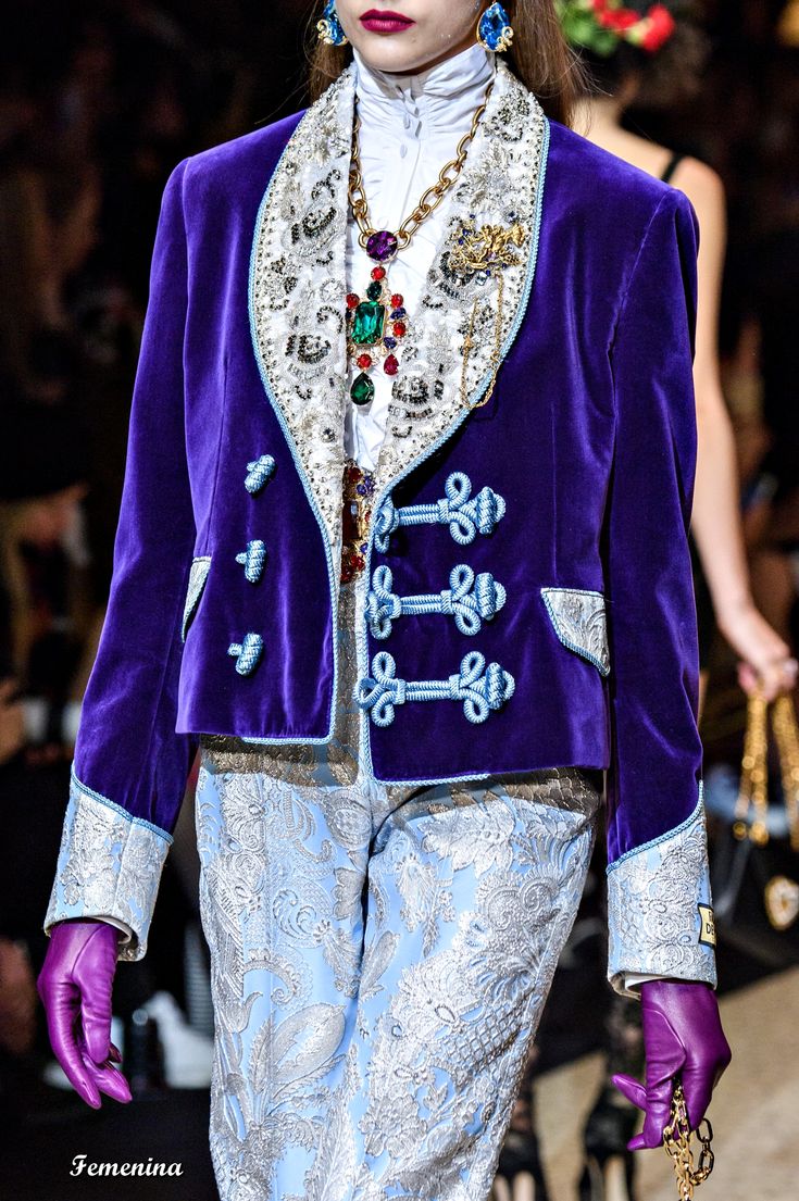 closeup shot of a model on a fashion runway, showcasing a striking outfit. the model is wearing a vibrant purple velvet jacket adorned with intricate silver embroidery, detailed buttons, and a lavishly embellished collar.  underneath, she wears light blue pants with a silver brocade pattern.  she is accessorized with a substantial statement necklace featuring large gemstones (emerald green and purple) and gold chains, matching earrings, and purple leather gloves. the overall style is opulent and luxurious, characteristic of highfashion runway presentations. the background is blurred, focusing attention on the model's clothing and accessories.  the image likely captures a moment from a dolce & gabbana fashion show.