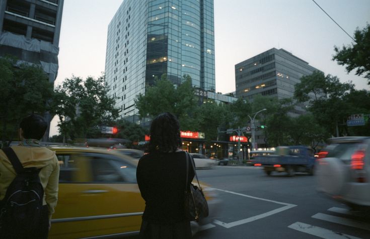 the image depicts a city street scene at dusk or twilight.  the setting appears to be an asian city, judging by the architecture.  two prominent skyscrapers dominate the background, with several other buildings lining the street.  the foreground shows a busy intersection with cars, including a yellow taxi cab, moving through a crosswalk.  a woman with a backpack stands near the curb, seemingly waiting to cross the street. the overall atmosphere is one of urban hustle and bustle, captured with a slight motion blur, giving a sense of movement and energy.  the light is soft, indicating the time of day is late afternoon or early evening.  trees line the streets, providing a touch of greenery amidst the urban landscape.