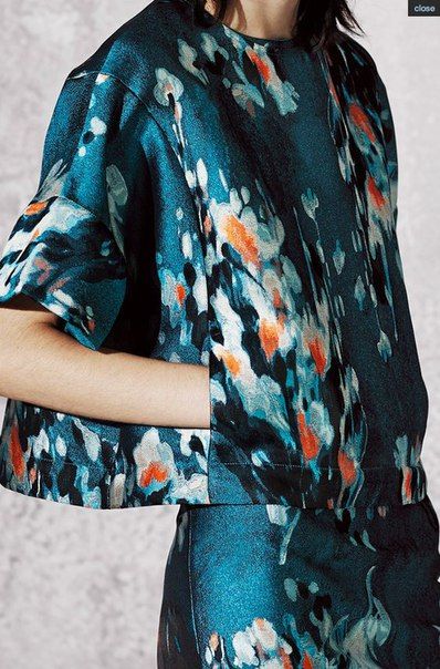 closeup shot of a woman's torso and waist wearing a teal twopiece outfit. the top and skirt are made of a teal fabric with an abstract floral print resembling painted splatter. the print features shades of teal, blue, white, and orange. the top is cropped and has short sleeves. the overall style is contemporary and stylish. the background is a blurry, neutral gray, making the outfit the focal point of the image. the image seems to be a fashion photograph, showcasing the details of the clothing.