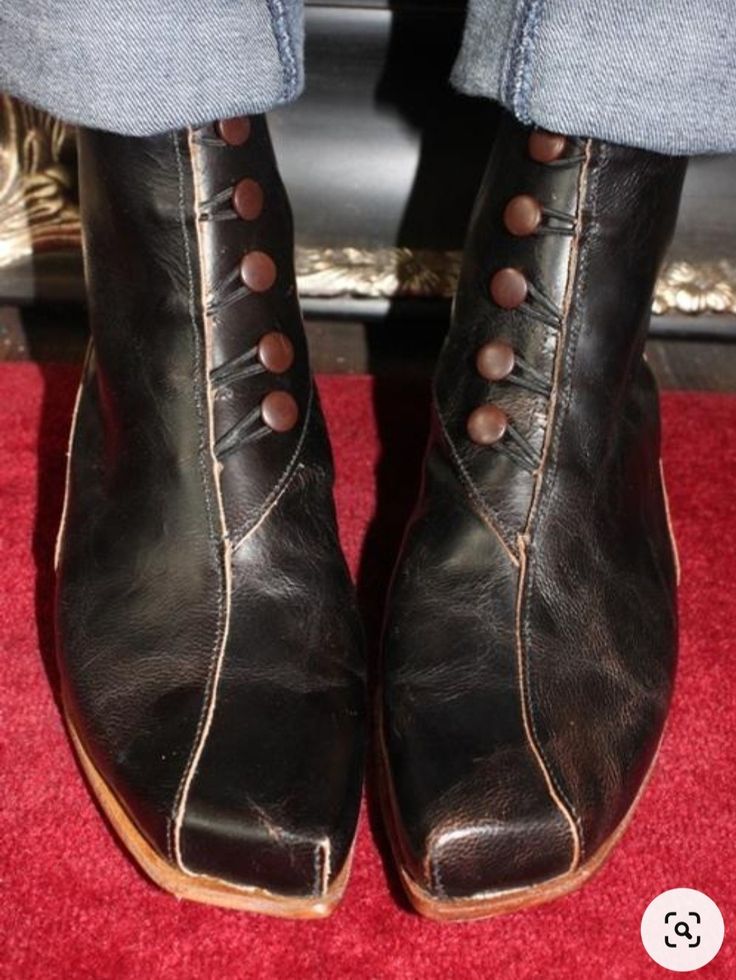 a closeup shot focuses on a pair of unique, black leather boots. the boots have a square toe, and are fastened with a series of dark brown buttons running up the shaft. the leather has a slightly distressed or vintage look. the boots are worn over jeans, and rest on a red carpet or surface. the overall style is retro or vintageinspired. the craftsmanship appears to be high quality, possibly handmade.